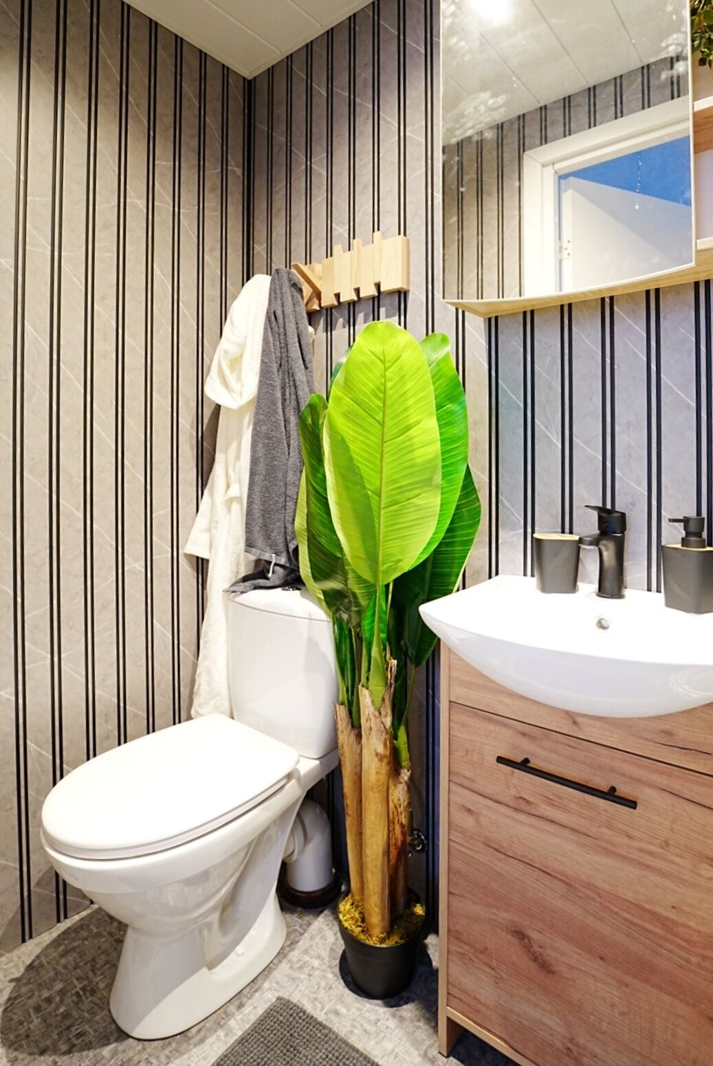 A bathroom with a striped wall, a white toilet, a wooden cabinet, and a large green plant.