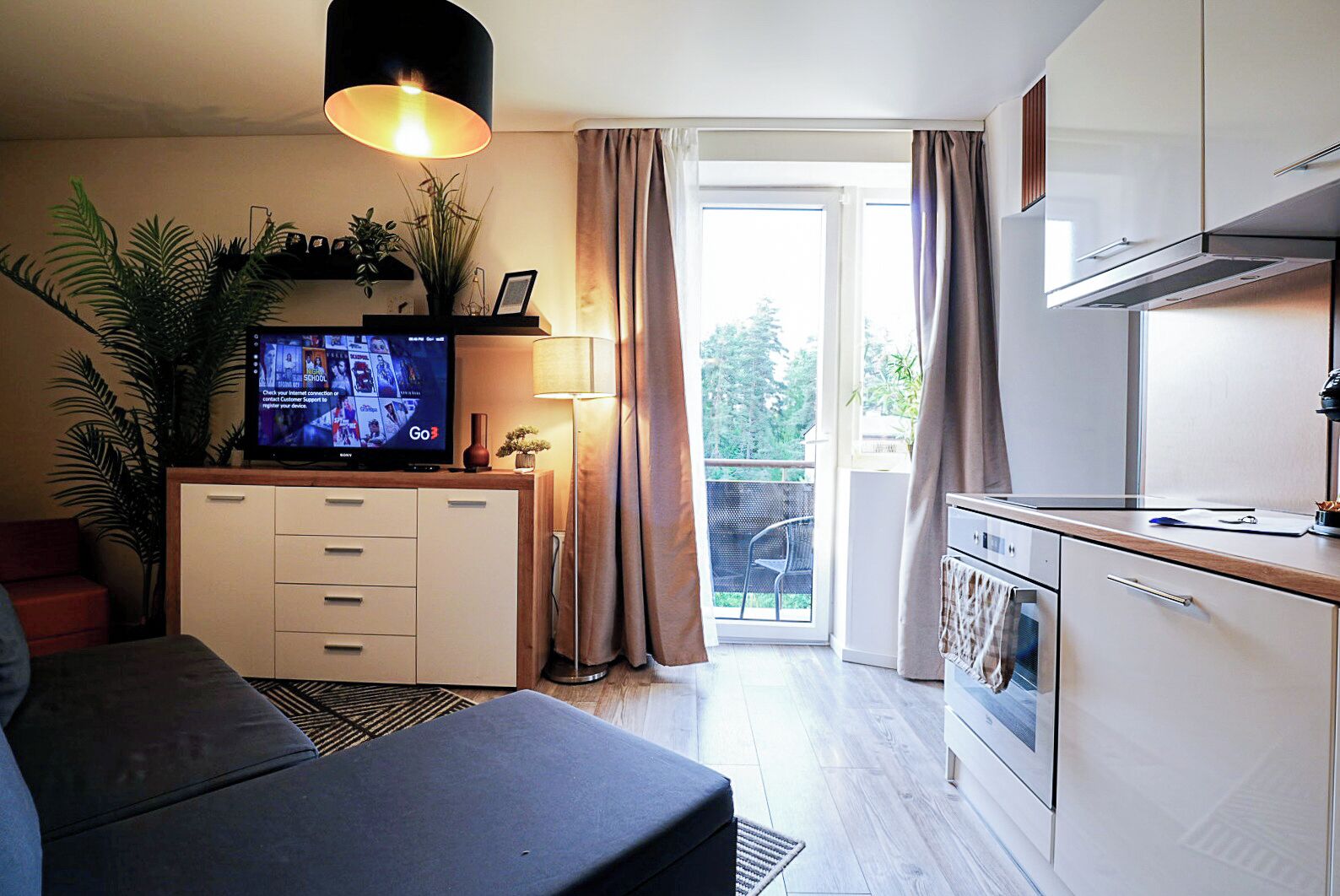 Ett modernt vardagsrum med en platt-TV, en soffa, en matplats med kök och en balkong med gardiner.
