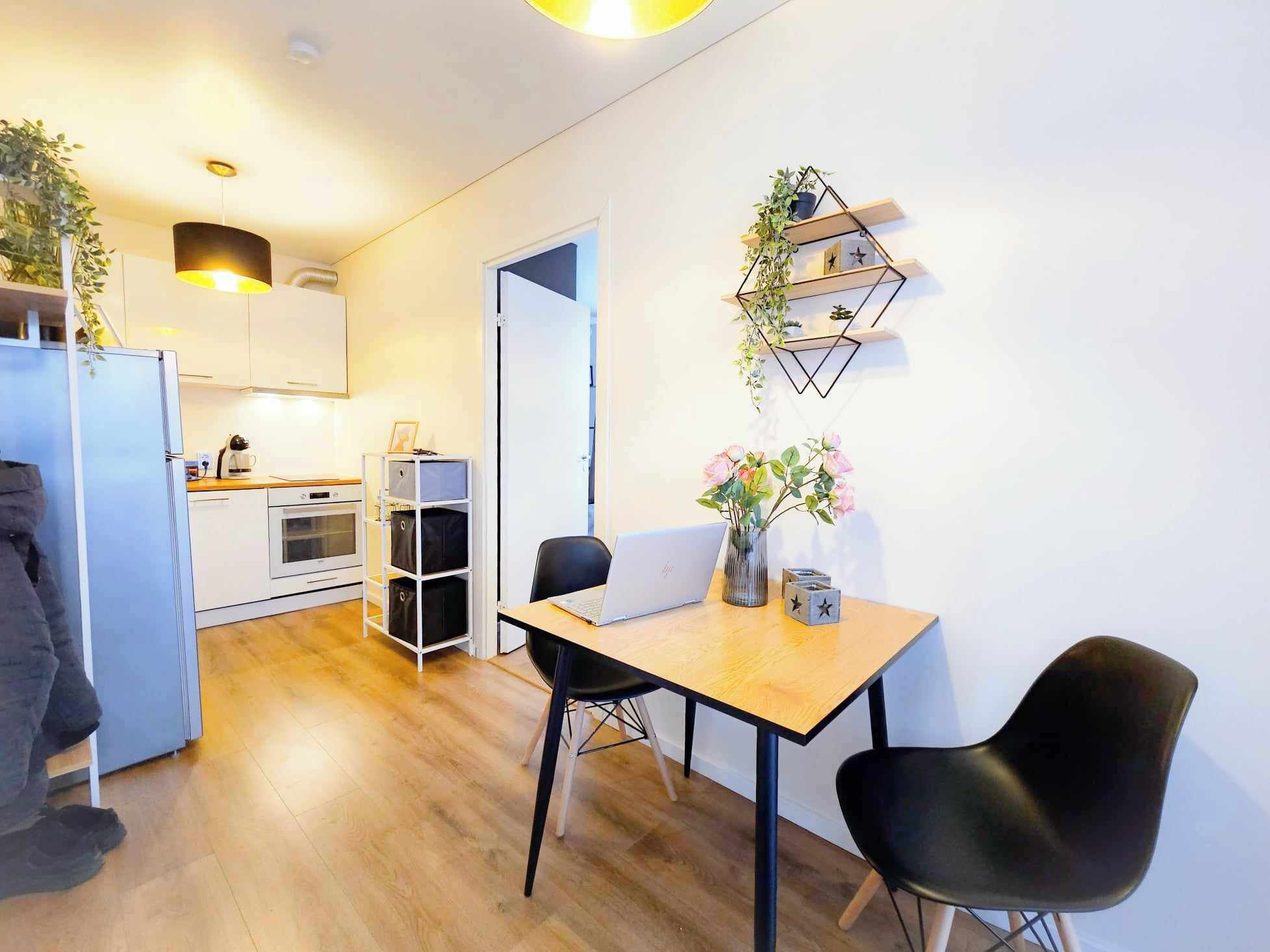 Una cocina moderna con armarios blancos, encimera de madera y horno empotrado. Hay una zona de comedor con mesa de madera, dos sillas negras y un ordenador portátil. Una planta está en la pared y una lámpara colgante ilumina la mesa.
