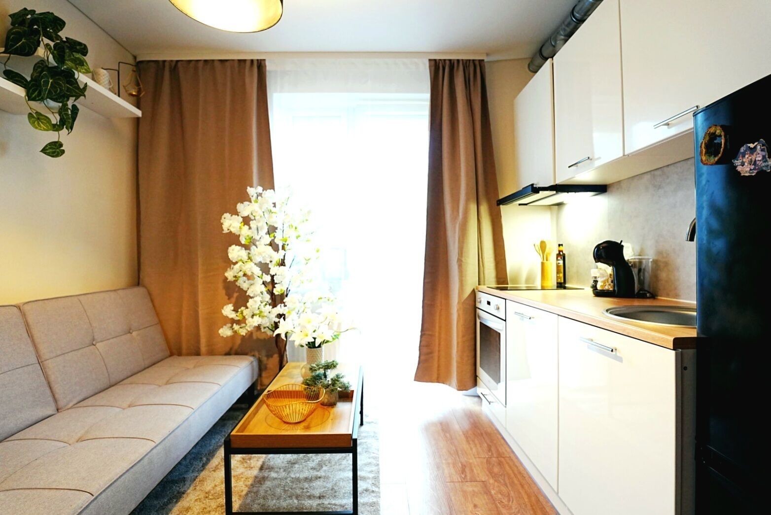 Ein kompakter Wohnbereich mit Sofa, Couchtisch, Küchenzeile mit Kühlschrank und einem Fenster mit Vorhängen.