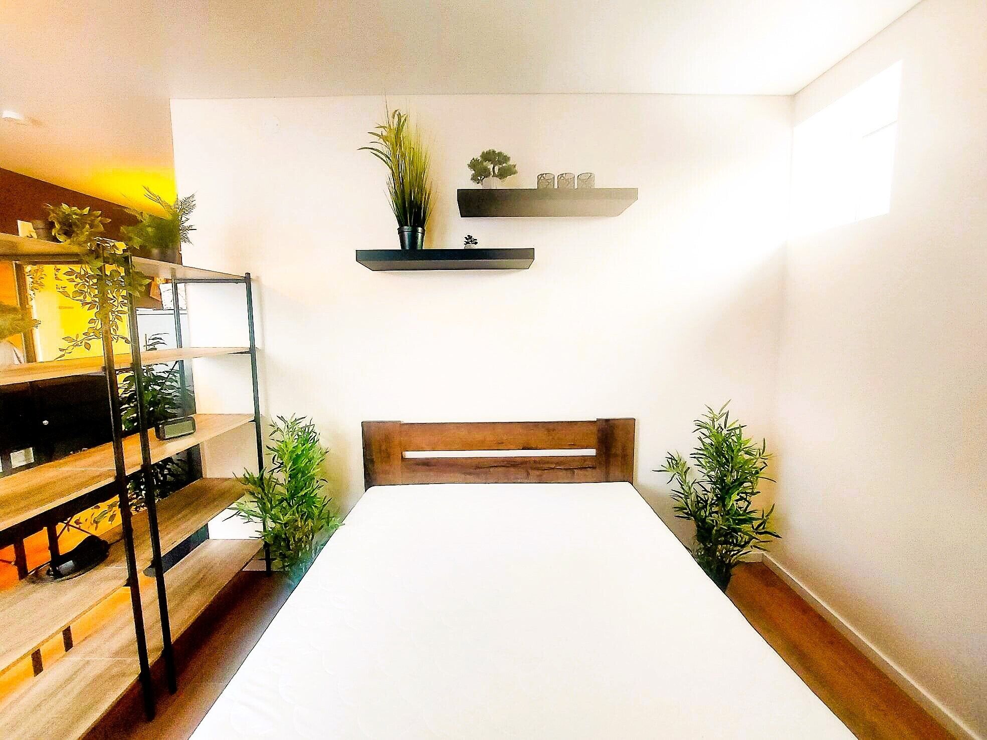 Una habitación pequeña y luminosa con una cama de madera, un escritorio con silla y un estante con objetos decorativos.