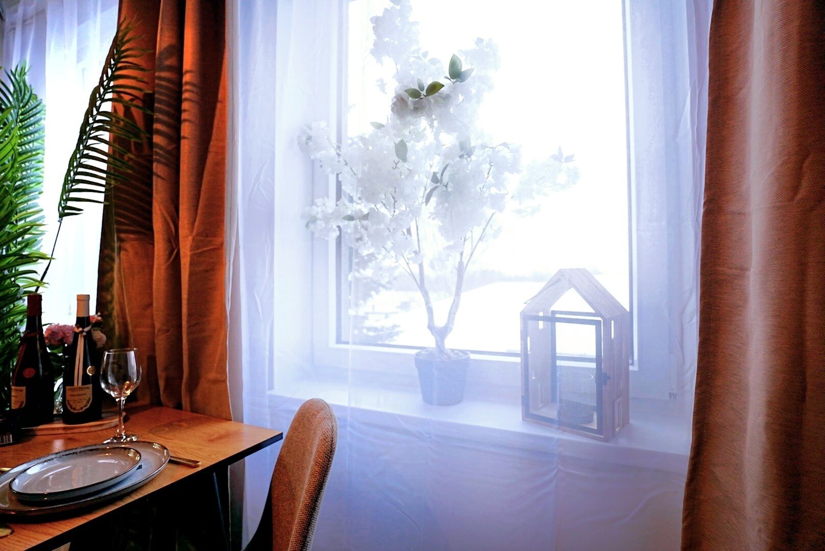 Uma área de jantar com uma mesa posta para dois, uma planta em vaso e uma janela com vista para uma casa branca e vegetação.