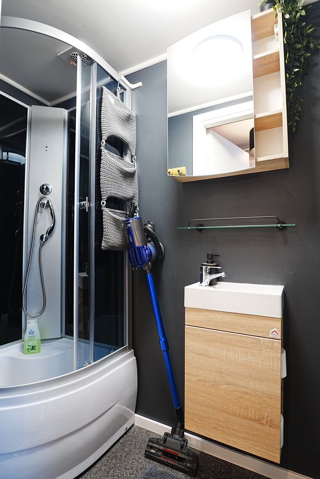 Un bany amb una cabina de dutxa de vidre, un aspirador blau, un lavabo blanc i un armari de fusta.