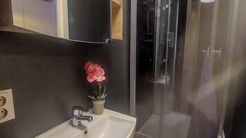 Um banheiro com pia branca, espelho, box de chuveiro e um vaso com planta.