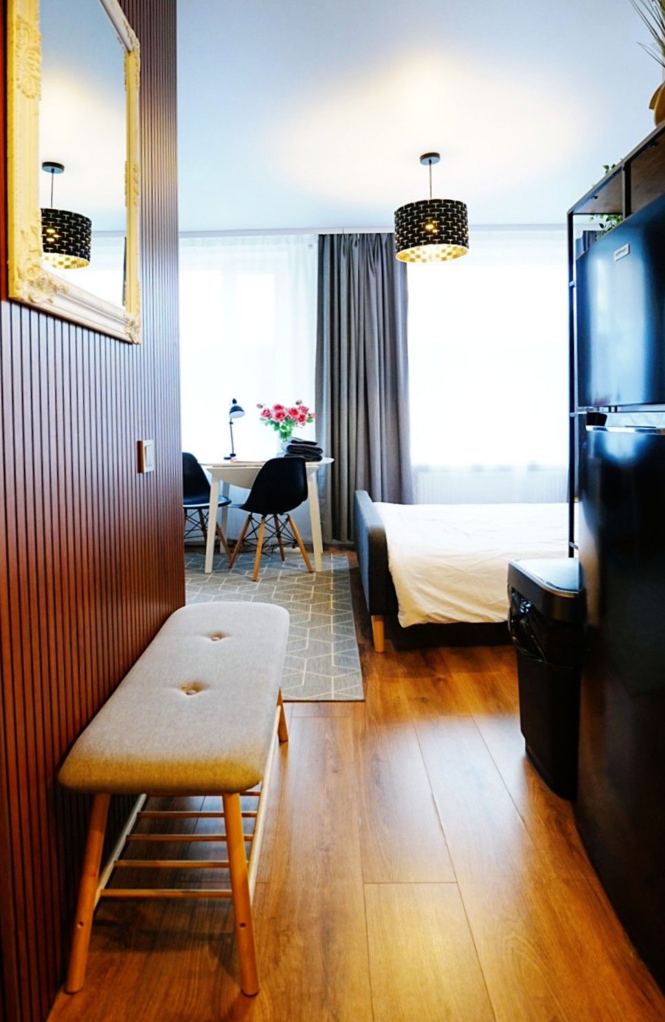 Habitación de hotel compacta con una banca de madera, una cama, una mesa de comedor con sillas, un televisor y un ventanal grande con cortinas.