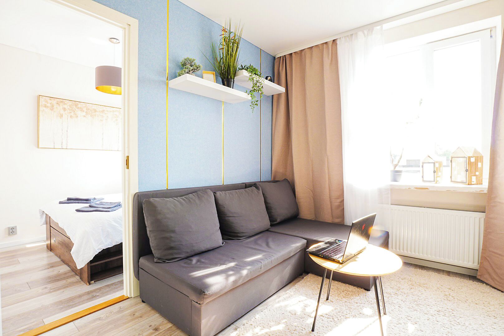 Una sala de estar moderna con un sofá gris, una mesita redonda con un ordenador portátil, una ventana con cortinas y una estantería con plantas.