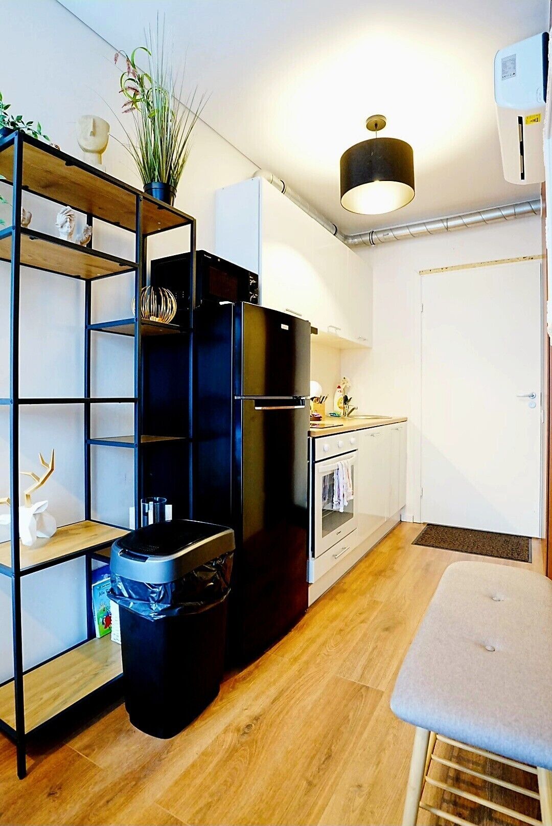 A compact kitchen with a built-in oven, a black refrigerator, a wooden shelf unit with decorative items, and a small dining table.