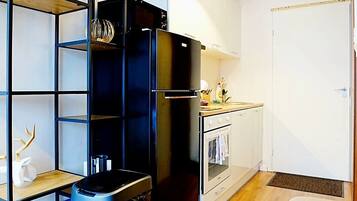 A compact kitchen with a built-in oven, a black refrigerator, a wooden shelf unit with decorative items, and a small dining table.