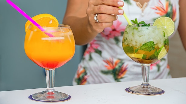 Zwei Cocktails in hohen Gläsern, mit Limettenscheibe und Minzblättern, und eine Person, die einen gelben Trinkhalm hält.