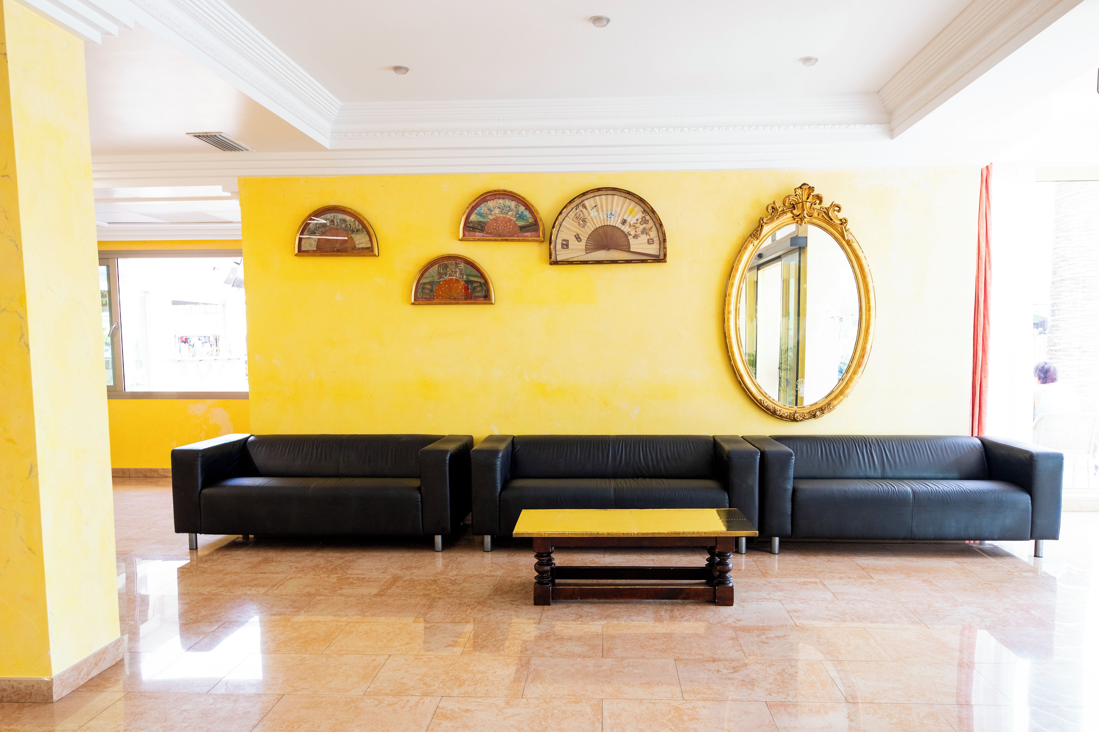 A lobby with a yellow wall, three black sofas, a wooden coffee table, and a large mirror.