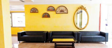 Un hall con pared amarilla, tres sofás negros, una mesa de centro de madera y un espejo grande.