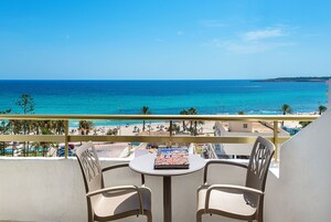 Un balcón con dos sillas y una mesa, con vista a una playa de aguas azules cristalinas y una isla distante.