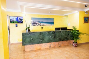 Un mostrador de recepción con encimera de mármol, una fotografía enmarcada de una playa, una planta en maceta y una cámara de seguridad en el techo.