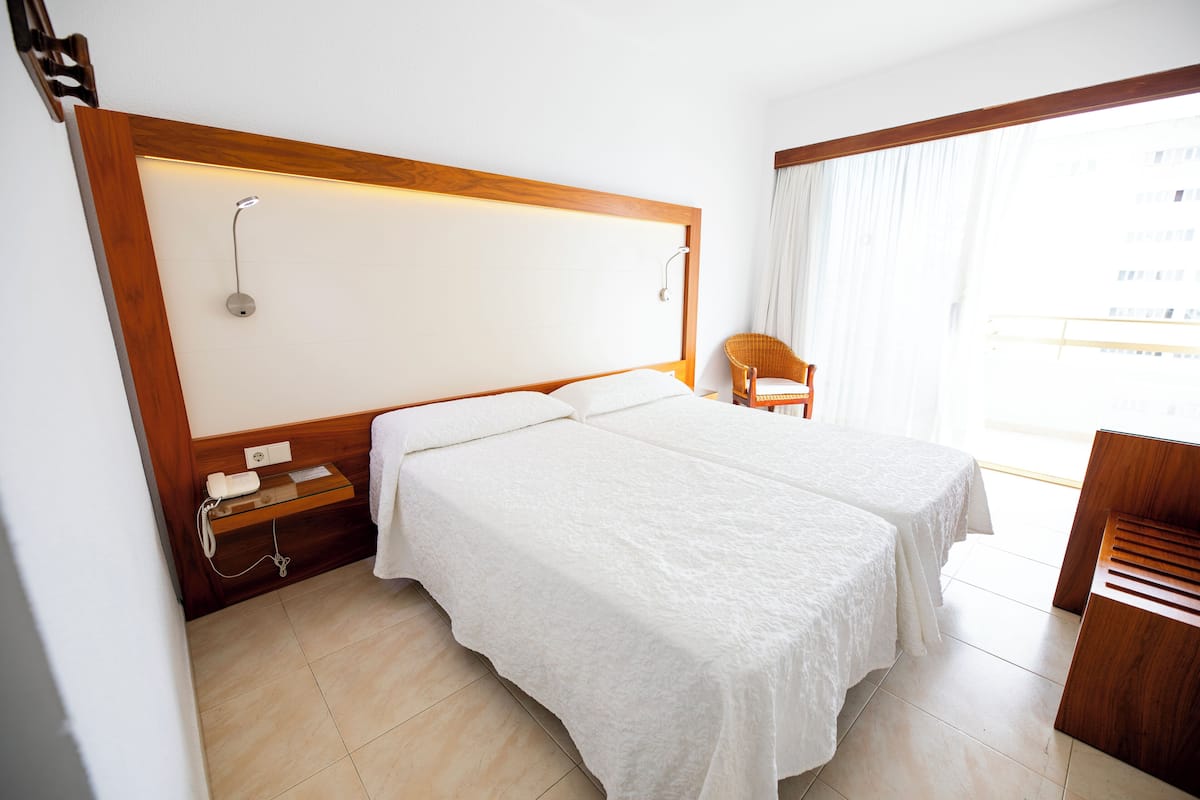 A hotel room with two beds, a wooden headboard, a small wooden side table, a chair, and a large window with curtains.