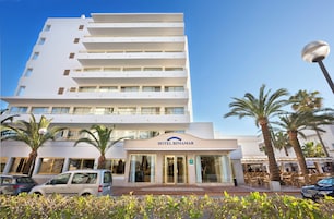 Un hotel de varios pisos con una entrada destacada, palmeras y un cielo azul despejado.