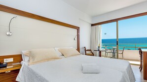 A hotel room with a large bed, a bedside table, a chair, a small table with a bottle, and a view of the sea.