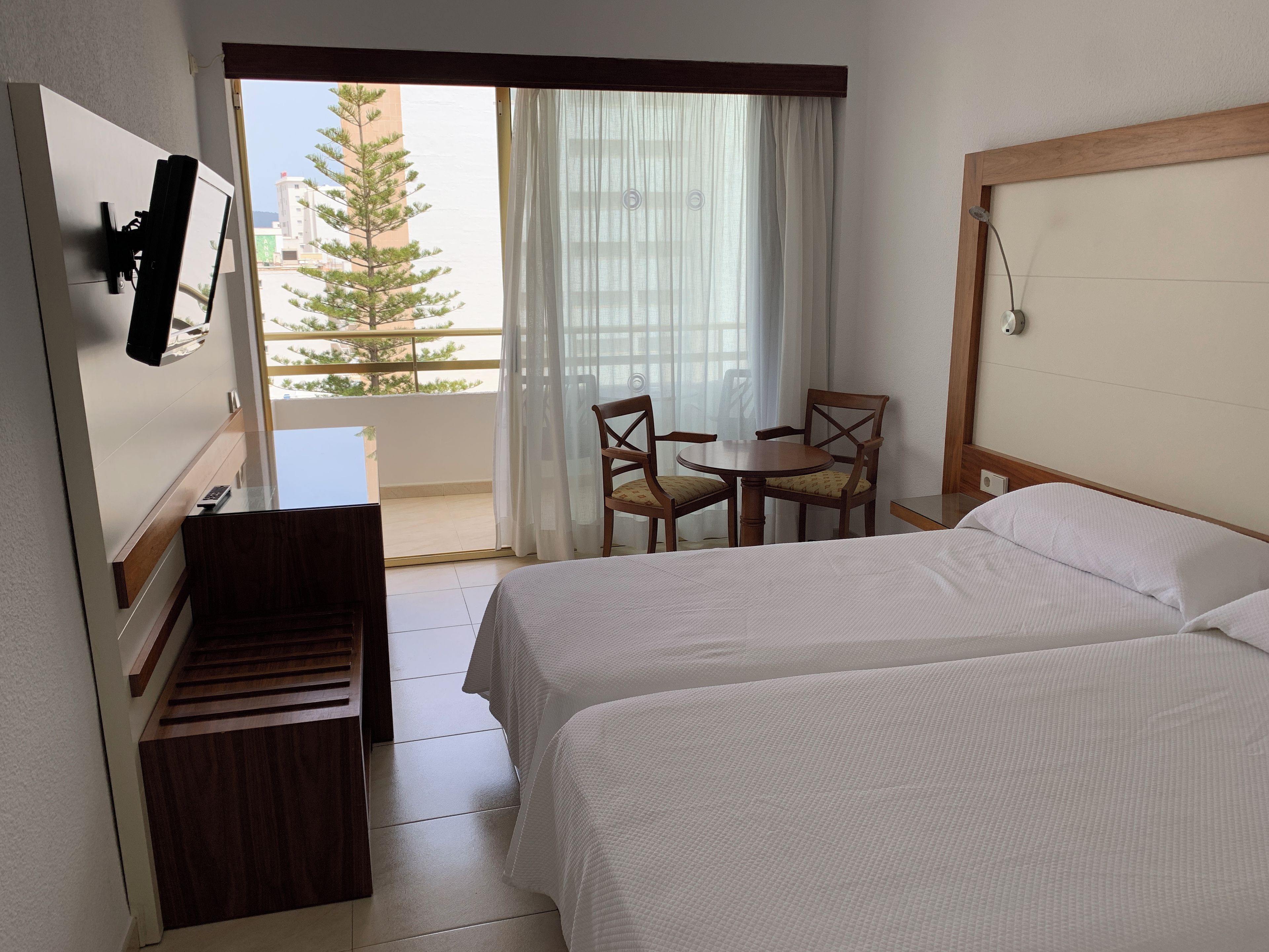 A hotel room with two beds, a desk, a chair, a TV, and a balcony with a view of buildings and trees.