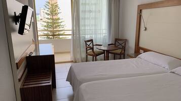 A hotel room with two beds, a desk, a chair, a TV, and a balcony with a view of buildings and trees.