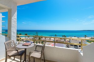 Un balcón con mesa y sillas, con vista a una playa y aguas azules cristalinas, y una costa distante.