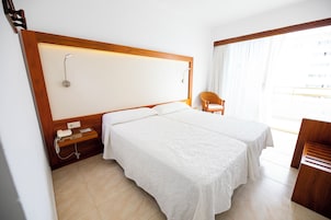 Habitación de hotel con dos camas, cabecero de madera, una mesita de noche pequeña de madera, una silla y un ventanal grande con cortinas.