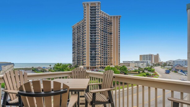 A balcony with outdoor furniture, a view of a tall building, and a road with vehicles. - The Ocean Bridge C34 - Beautiful Unit Across from Beach (Myrtle Beach)