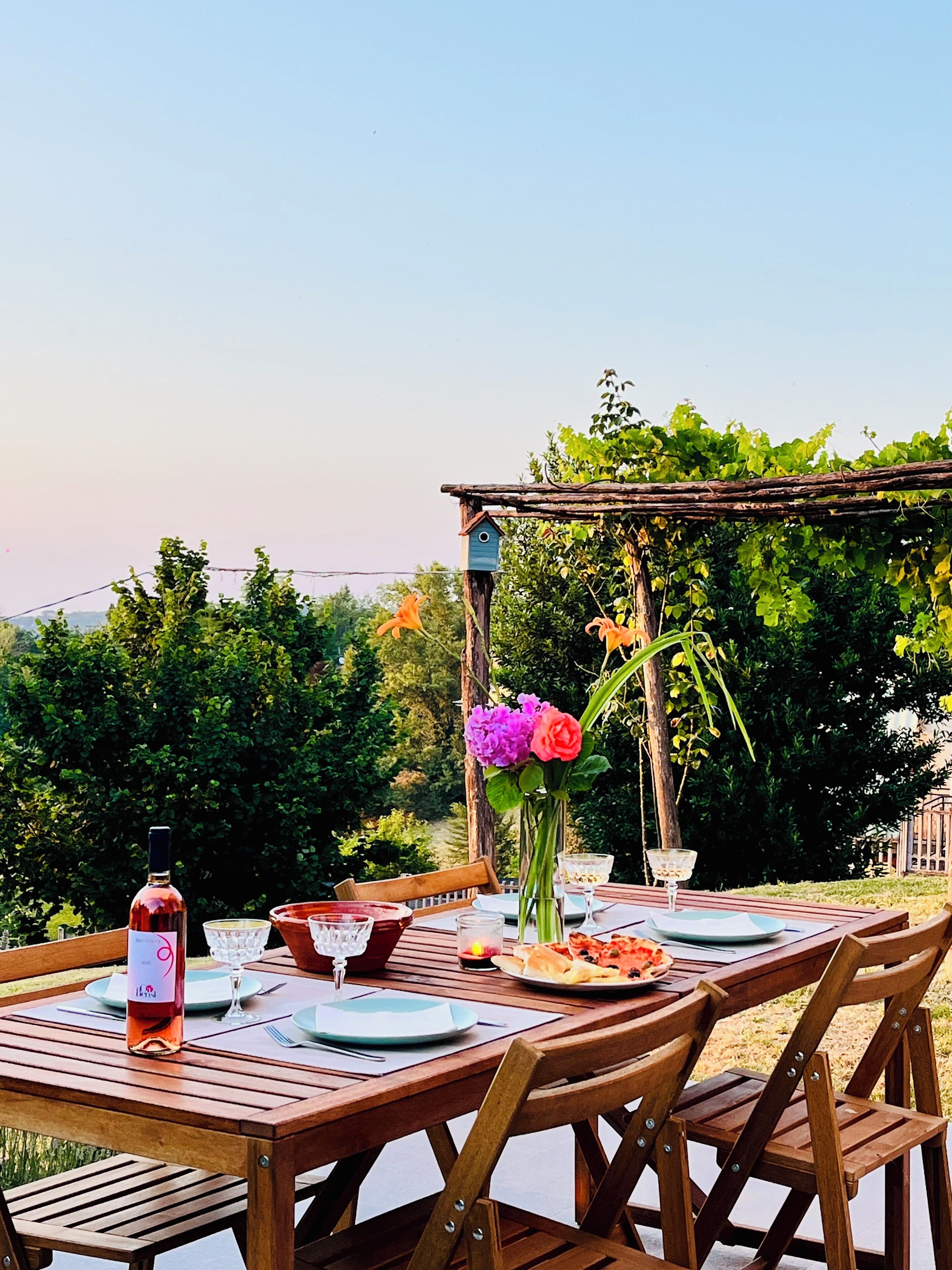 Un'area pranzo all'aperto con tavolo in legno apparecchiata per due, circondata da piante e una pergola.