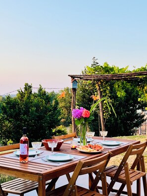 Un área de comedor al aire libre con una mesa de madera dispuesta para dos personas, rodeada de follaje y una pérgola.