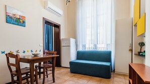 A compact living space with a dining table and chairs, a sofa, a small refrigerator, and a window with sheer curtains. - Grimaldi 8 Apartment by Wonderful Italy (Catania)