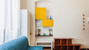 A room with a blue sofa, a white bench, a wooden table, and a yellow shelf with a clock and decorative items. - Grimaldi 8 Apartment by Wonderful Italy (Catania)