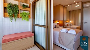 Une chambre d’hôtel moderne avec un banc en bois, une grande fenêtre avec des rideaux, un lit avec des serviettes soigneusement pliées et un miroir qui reflète la pièce.