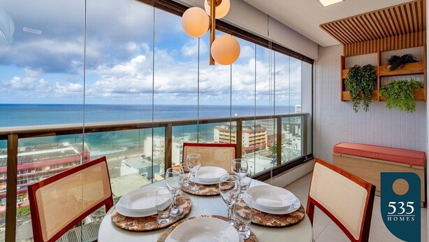Un coin repas avec une table dressée pour quatre personnes, de grandes fenêtres offrant une vue sur la ville et une étagère en bois ornée de plantes.
