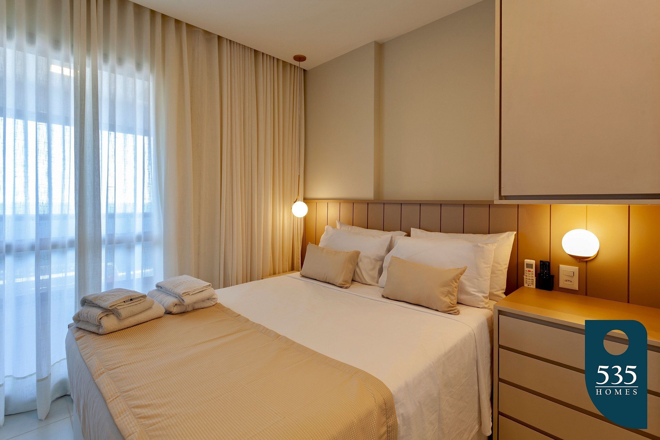 A hotel room with a bed, bedside tables, a nightstand, and a large window with curtains.