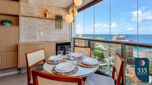 Un coin repas avec une table dressée pour six personnes, un luminaire suspendu et une vue sur l’océan grâce à de grandes fenêtres.