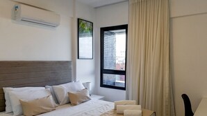 Una cama bien hecha con cabecera gris, ropa de cama blanca y una manta beige. Se ve una ventana con vistas a edificios y una cortina a la derecha.