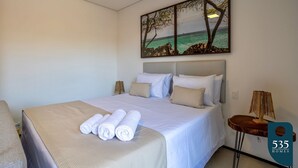 A hotel room with a large bed, white linens, a painting of a beach scene, and a small table with a lamp. - Apartment in Itacaré: Sun, Sea, and Comfort (Itacaré)