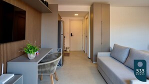 A modern living room with a sofa, a TV mounted on the wall, a dining area with a table and chairs, and a kitchen area with cabinets. - Apartment in Itacaré: Sun, Sea, and Comfort (Itacaré)