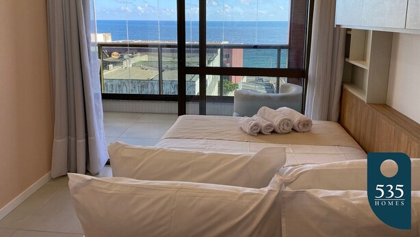 Une chambre d’hôtel dotée d’une grande fenêtre donnant sur la mer et les bâtiments, un lit avec des serviettes soigneusement pliées, et une armoire en bois.