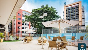 Un patio al aire libre con mobiliario de mimbre y sombrillas, un parque infantil y vistas a un edificio con fachada azul.