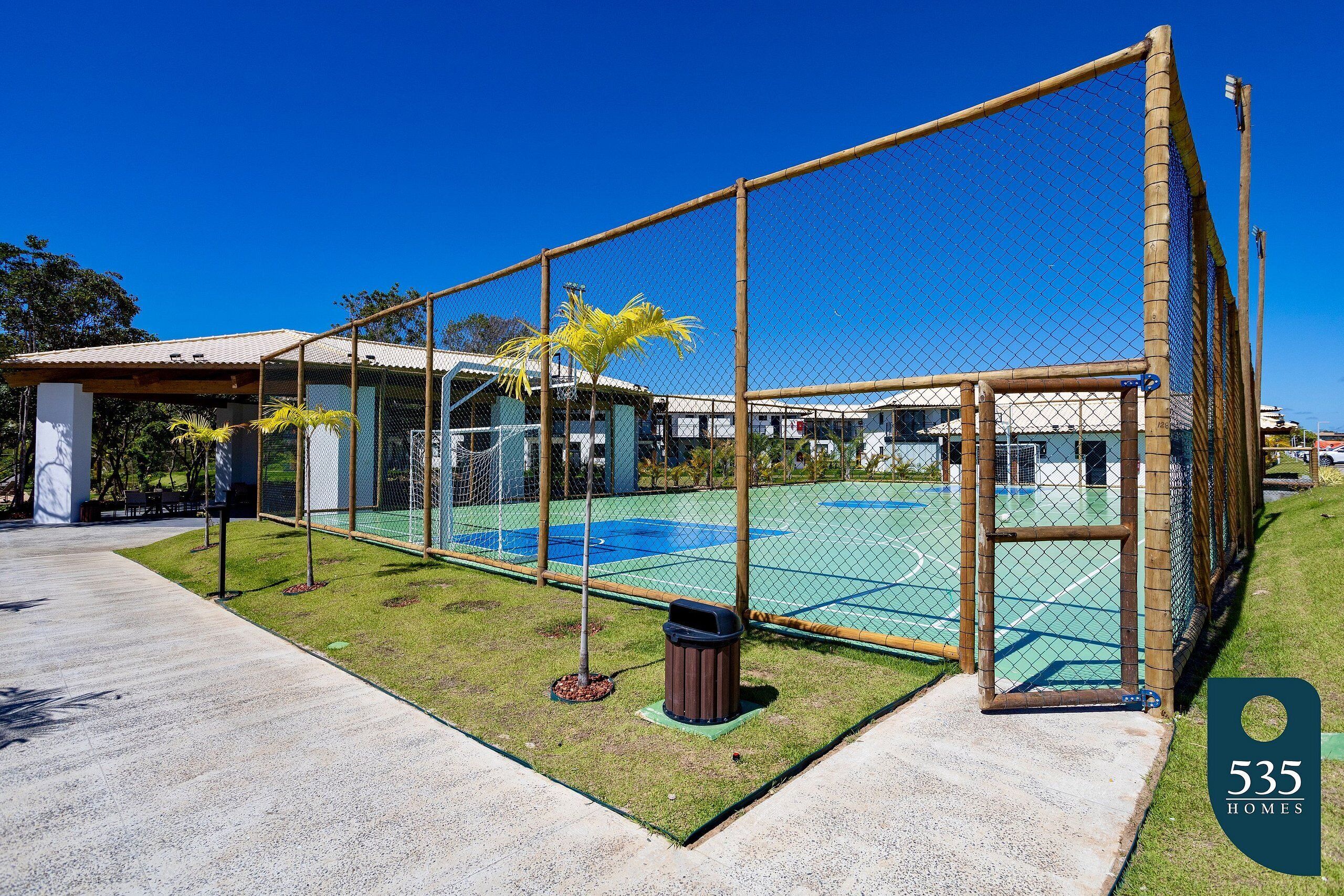 A fenced outdoor area with a basketball court, a trash bin, and a building with a tiled roof.