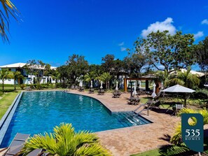 Une piscine entourée de verdure, avec des chaises longues et des parasols, sous un ciel bleu azur.