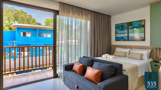 Une chambre d’hôtel moderne avec un balcon, un lit doté d’une tête de lit blanche, un canapé gris et un tableau accroché au mur.