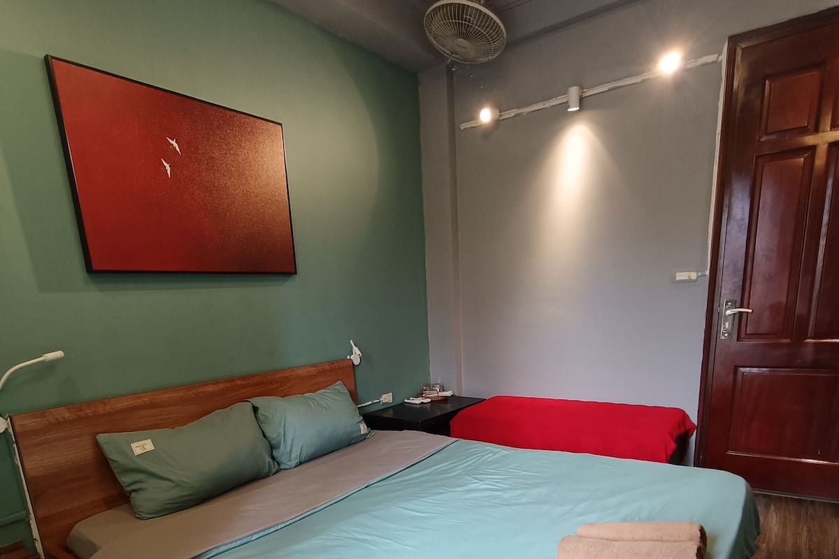 A bedroom with a wooden bed, a red pillow, a blue bedspread, a wooden floor, a wall clock, and a ceiling fan.