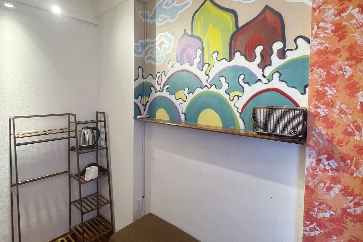 A room with a wooden bench, a metal shelf with a kettle, a coffee maker, and a colorful mural on the wall.