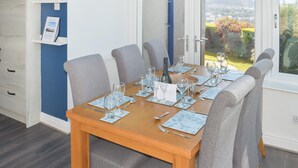 Ein Esszimmer mit einem Holztisch, gedeckt für sechs Personen, grau gepolsterten Stühlen und Blick auf einen Garten durch eine Glastür.