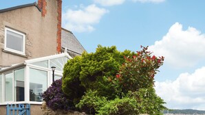 Ein Haus mit Ziegelkamin, Wintergarten und verschiedenen Pflanzen, darunter lila Blumen und ein Baum mit roten Blättern.