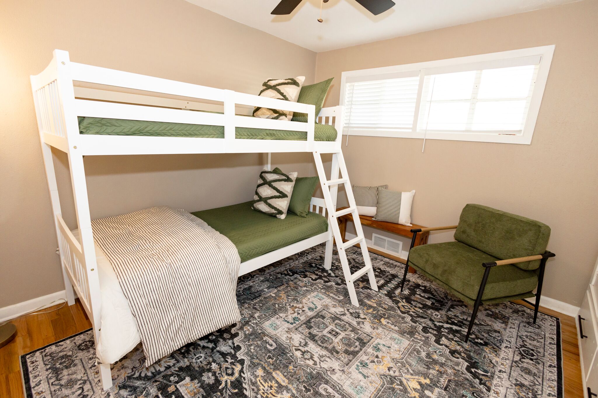 A bunk bed with a ladder, a green chair, and a patterned rug in a room with beige walls and a window.