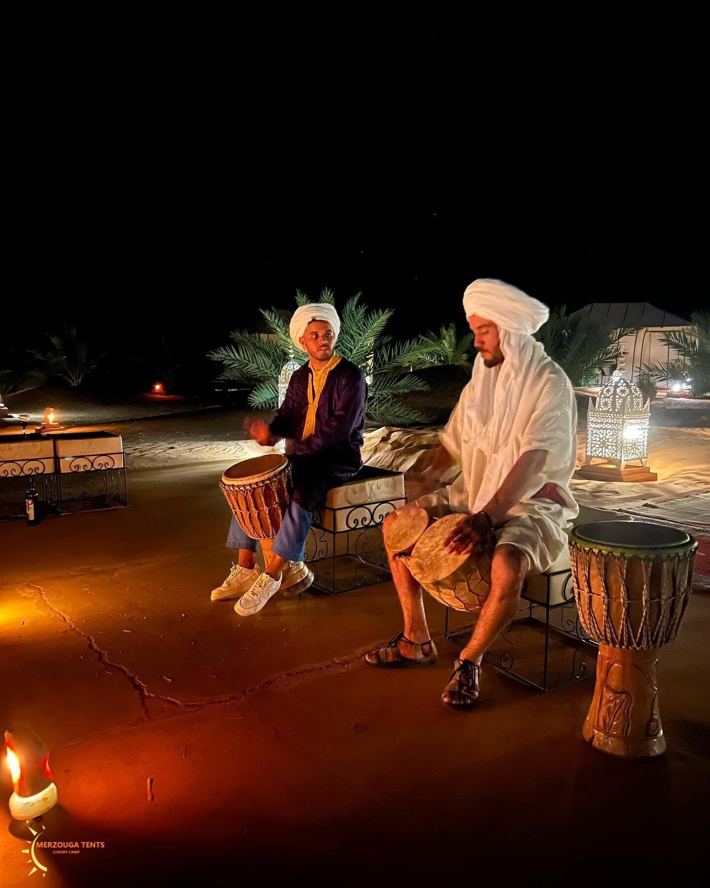 Two individuals playing drums outdoors at night, with a tent and decorative lanterns in the background.