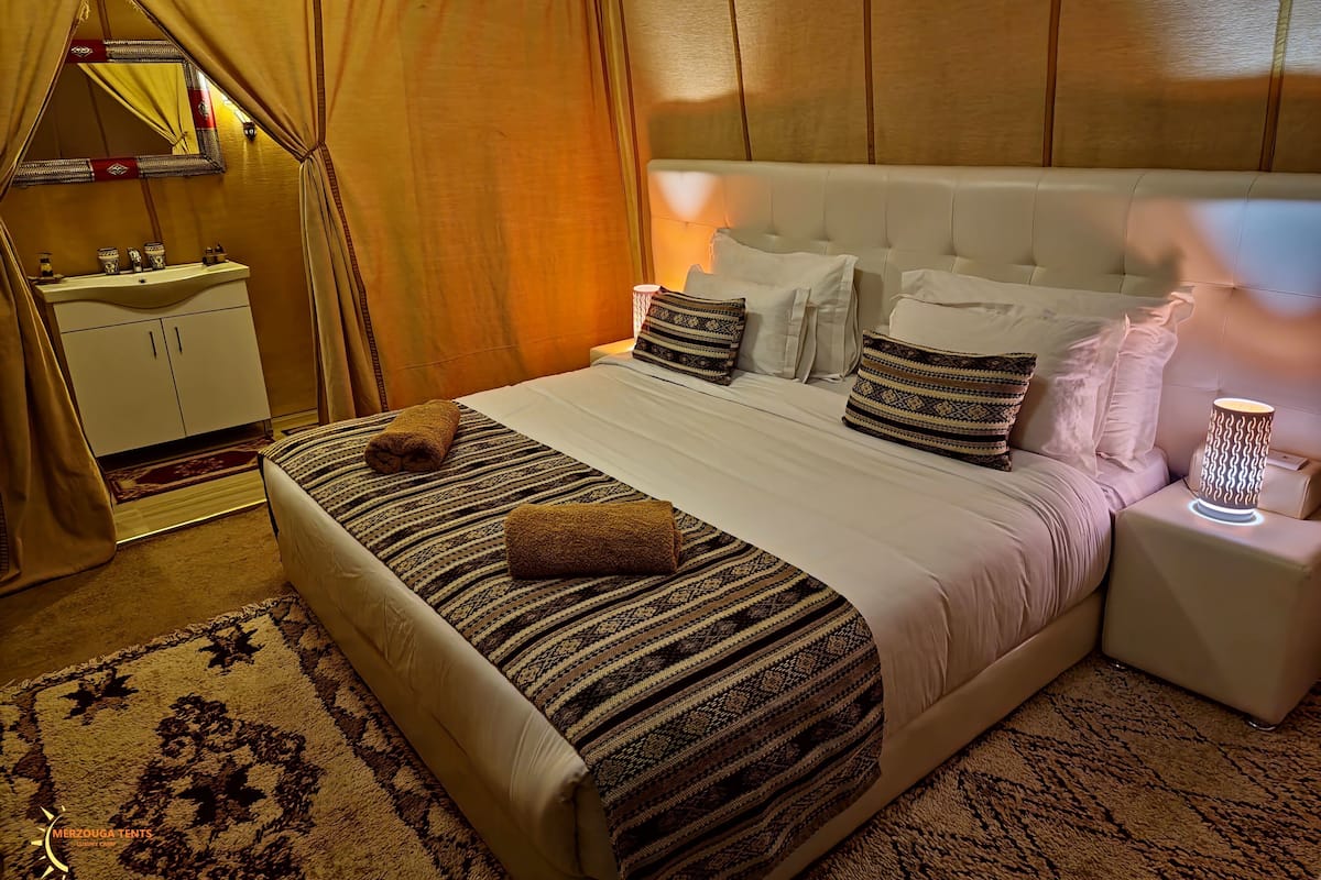Une chambre d’hôtel avec un grand lit, une table de chevet avec une lampe, un miroir, une coiffeuse avec un lavabo et un tapis à motifs.