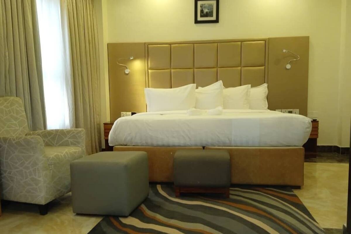 A hotel room with a large bed, two ottomans, a patterned armchair, and a striped area rug.