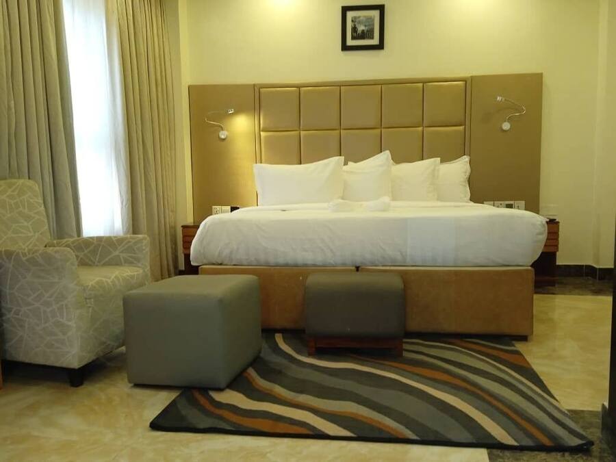 Une chambre d’hôtel avec un grand lit, deux poufs, un fauteuil à motifs et un tapis à rayures.